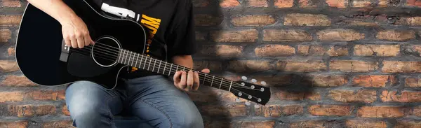 Music and sound - fragment man play a black acoustic guitar on a old brick wall background.