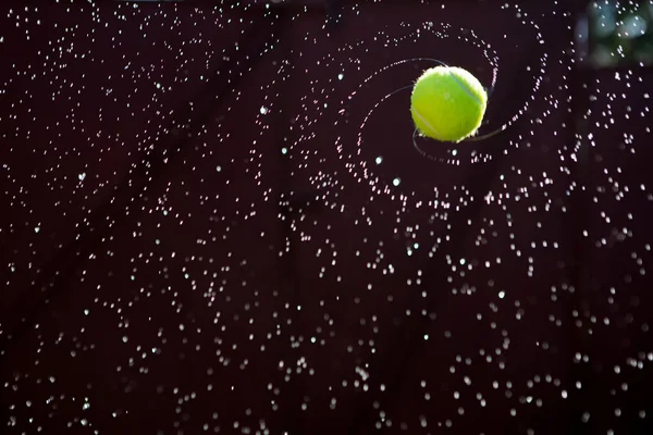 Nasser Tennisball, fotografiert beim Fliegen und Drehen bei Sonnenuntergang an einem warmen Sommerabend. Spritzwasser fliegt in verschiedene Richtungen — Stockfoto