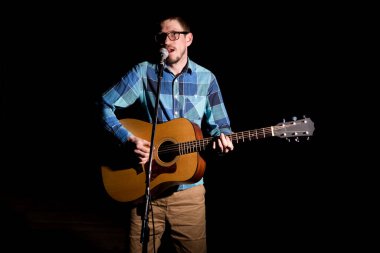 Akustik gitar çalıp şarkı söyleyen ekose gömlekli genç bir müzisyen..