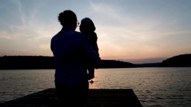 Anne ve bebek gün batımında iskelede. Sıcak, güneşli bir yaz gününde dağlarda. Siluetler.