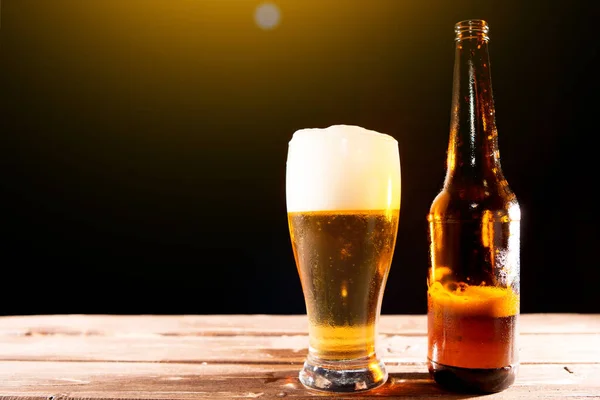 Birra fresca e gustosa nella calda giornata estiva. Bottiglia e bicchiere di birra su un tavolo di legno su sfondo nero e spazio copia — Foto Stock