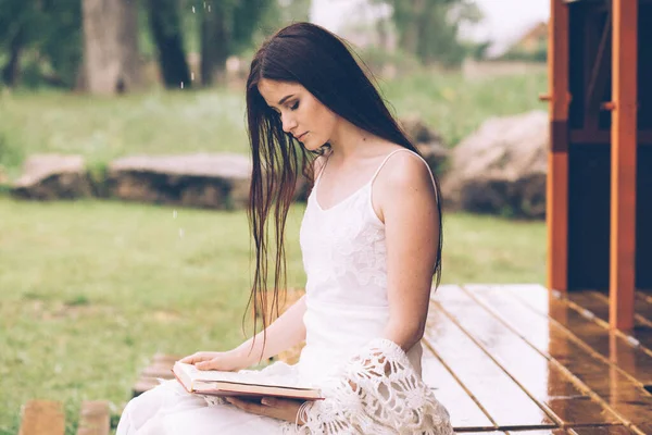 Ładna kobieta w białej sukience czyta książkę w pochmurnej naturze. Dziewczyna siedzi na tarasie lub na ganku — Zdjęcie stockowe