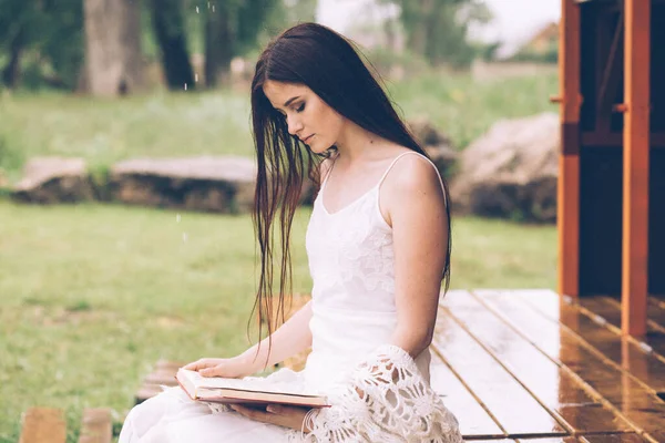 Hübsche Frau im weißen Kleid liest Buch in der bewölkten Natur. Mädchen sitzt auf der Terrasse oder auf der Veranda — Stockfoto