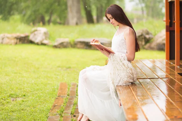 Hübsche Frau im weißen Kleid liest Buch in der bewölkten Natur. Mädchen sitzt auf der Terrasse oder auf der Veranda — Stockfoto