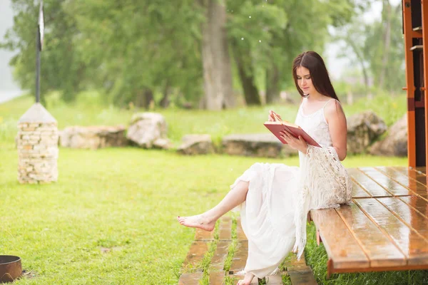 Ładna kobieta w białej sukience czyta książkę w pochmurnej naturze. Dziewczyna siedzi na tarasie lub na ganku — Zdjęcie stockowe