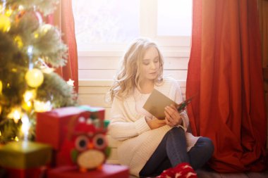Genç ve güzel bir kadın Noel ağacının yanında oturmuş Noel kartı okuyor.