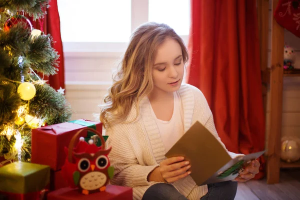 Joven hermosa mujer sentada en el suelo cerca del árbol de Navidad y leyendo la postal de Navidad — Foto de Stock