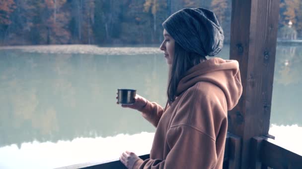 La chica está descansando en una caminata. Bebe té o café de un termo en la orilla de un lago de montaña. Vídeo en cámara lenta — Vídeos de Stock