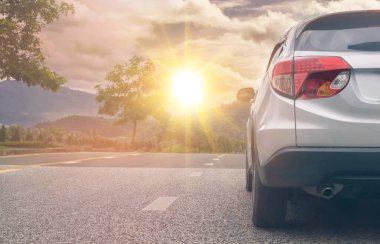 Gümüş renkli SUV arabası asfalt yolda gün batımı ya da gün doğumuyla birlikte turuncu gökyüzü dağı ve ağaçlarla birlikte bir doğa konseptiyle araba ile seyahat eder.