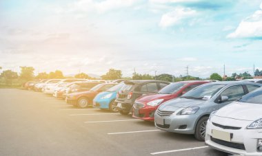 Güneşli bir günde, büyük asfalt otoparkta beyaz bulutlu ve mavi gökyüzü arka planlı bir araba parkı. Açık hava otoparkı, taze ozon ve yeşil ulaşım ve teknoloji ortamı.