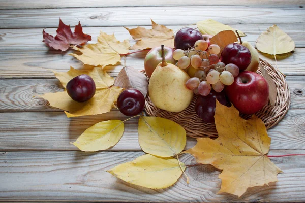 Herbststilleben. Auf dem Korbteller und dem Holztisch liegen verschiedene Früchte. Herbstgelbe Blätter. — Stockfoto