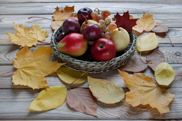 様々な果物を持つ秋の静物が、ウィッカープレートに横たわっています。秋のカエデやその他の葉. — ストック写真