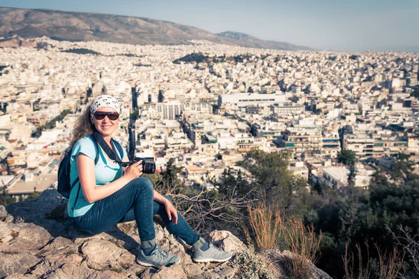 かわいい若い女性は カメラ ギリシャで上からアテネの都市景観と笑顔の写真を撮る 女性旅行者は アテネ市街を見渡せる頂上に座っています アテネの丘が観光スポットです — ストック写真