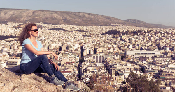 Young woman is on background of urban landscape of Athens, Greece, Europe. Adult pretty girl tourist relaxes on hilltop overlooking Athens city in summer. Concept of travel and vacation in Athens. 