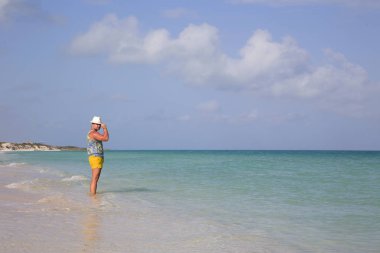 Genç bir adam denizde duruyor, şapkasını tutuyor ve bir kenara bakıyor. Yatay.