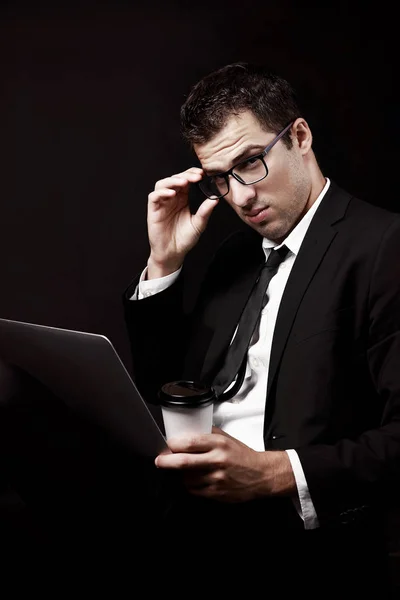 Brutal guy in a black suit with a coffee and a laptop, glasses. Black background. Coffee with you.  Manager.