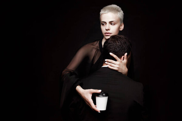 Girl with short hair hugs a guy with coffee on a black background. Passion. Creative advertising.