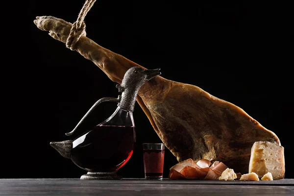 A carafe of wine in the form of a duck, a glass of wine, a leg of Parma ham and cheese. Black background.