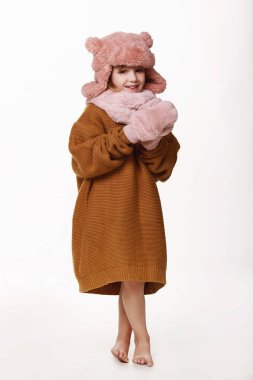 Kazaklı, şapkalı, eldivenli iyileşen kız yerde yalınayak yürüyor. Hastalık, soğuk algınlığı, grip, kış, sonbahar. Dokunulmazlık. Stüdyoda beyaz bir arkaplanda.