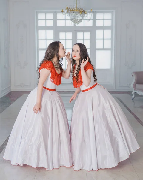Two Beautiful woman in white and red medieval dresses with crinoline whisper in the hall
