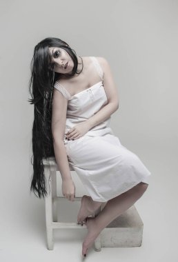 Siyah uzun saçlı, beyaz gömlekli gizemli hayalet kadın gri arka planda, sandalyede oturuyor.