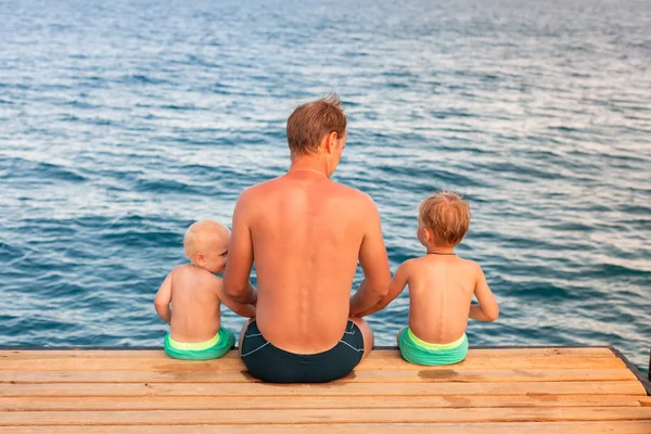 Padre e hijos balancean sus piernas desde el muelle de madera — Foto de Stock