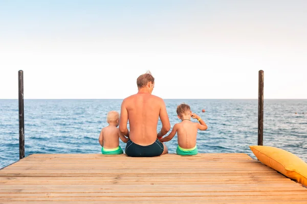 Padre e hijos balancean sus piernas desde el muelle de madera — Foto de Stock