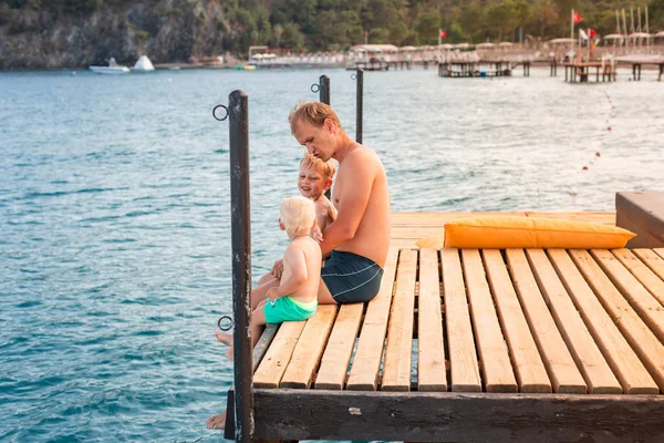 Padre e hijos balancean sus piernas desde el muelle de madera — Foto de Stock