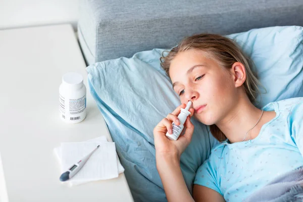 高熱と頭痛のある病気の十代の少女は 彼女の鼻に薬を噴霧ベッドに横たわっています コロナウイルスの流行時に家にいて病気になったら — ストック写真