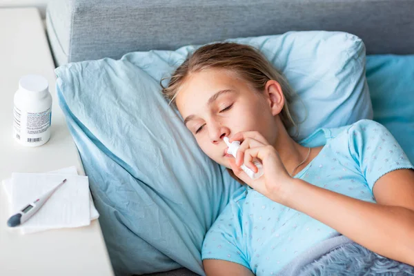 高熱と頭痛のある病気の十代の少女は 彼女の鼻に薬を噴霧ベッドに横たわっています コロナウイルスの流行時に家にいて病気になったら — ストック写真