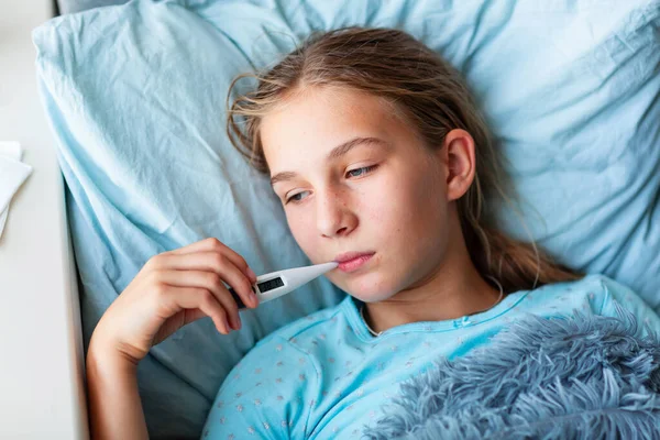 高熱と頭痛のある病気の十代の少女はベッドに横たわって 彼女の口の中で温度計を保持しています コロナウイルスの流行時に家にいて病気になったら — ストック写真