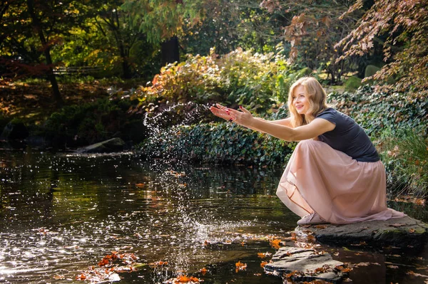 年轻女子玩水 坐在秋天城市公园湖边的石头上 — 图库照片