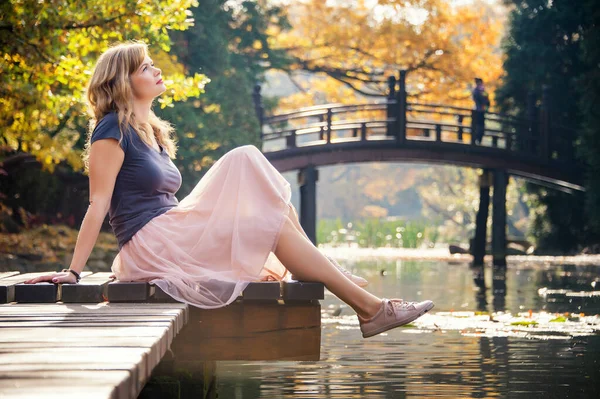 在秋天的城市公园里 梦中的年轻女子坐在湖边的一个木制码头上 — 图库照片
