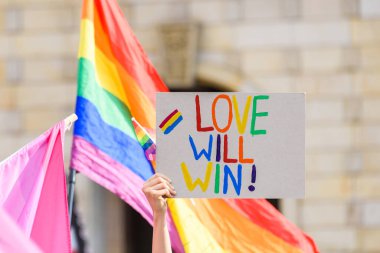 İnsan eli, üzerinde aşk yazan bir poster tutar ve arka planda LGBT gökkuşağı bayrağı olur.