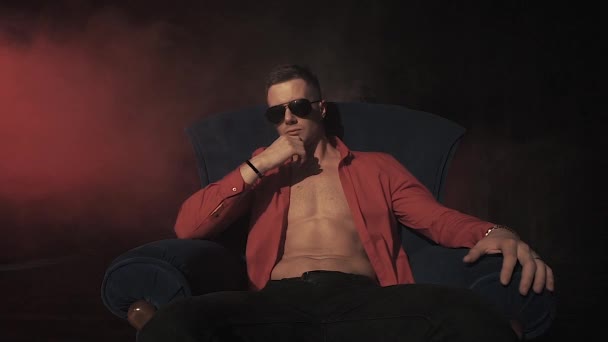 Sexy hombre en rojo camisa sentado en silla en humo fondo — Vídeos de Stock