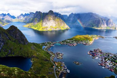 Göz kamaştırıcı dağların güneşli panoramik manzarası ve Reinebringen tepesinden Norveç 'in Lofoten adalarındaki Reine köyü. Gökkuşağı ve engebeli tepeleri olan manzaralı bir manzara.
