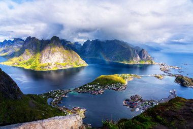 Göz kamaştırıcı dağların güneşli panoramik manzarası ve Reinebringen tepesinden Norveç 'in Lofoten adalarındaki Reine köyü. Gökkuşağı ve engebeli tepeleri olan manzaralı bir manzara.