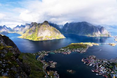 Göz kamaştırıcı dağların güneşli panoramik manzarası ve Reinebringen tepesinden Norveç 'in Lofoten adalarındaki Reine köyü. Gökkuşağı ve engebeli tepeleri olan manzaralı bir manzara.