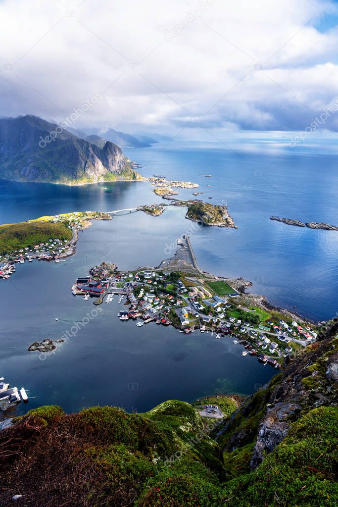 Sunny aerial panoramic view on stunning mountains and village of Reine in Lofoten islands, Norway from Reinebringen ridge. Scenic vista with a rainbow and rugged peaks.