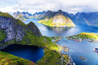 Göz kamaştırıcı dağların güneşli panoramik manzarası ve Reinebringen tepesinden Norveç 'in Lofoten adalarındaki Reine köyü. Gökkuşağı ve engebeli tepeleri olan manzaralı bir manzara.