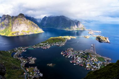 Göz kamaştırıcı dağların güneşli panoramik manzarası ve Reinebringen tepesinden Norveç 'in Lofoten adalarındaki Reine köyü. Gökkuşağı ve engebeli tepeleri olan manzaralı bir manzara.