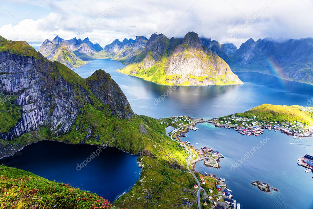 Sunny aerial panoramic view on stunning mountains and village of Reine in Lofoten islands, Norway from Reinebringen ridge. Scenic vista with a rainbow and rugged peaks.