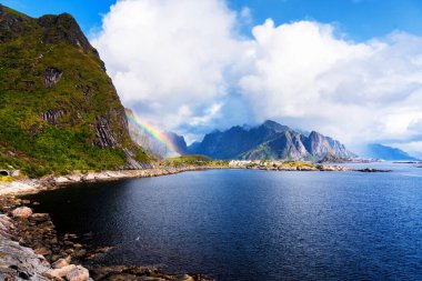 Göz kamaştırıcı dağların güneşli panoramik manzarası ve Reinebringen tepesinden Norveç 'in Lofoten adalarındaki Reine köyü. Gökkuşağı ve engebeli tepeleri olan manzaralı bir manzara.