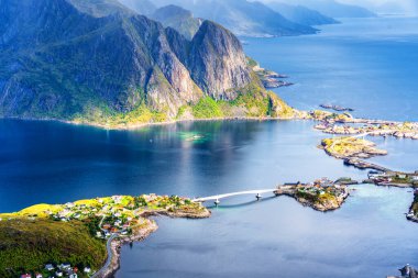 Göz kamaştırıcı dağların güneşli panoramik manzarası ve Reinebringen tepesinden Norveç 'in Lofoten adalarındaki Reine köyü. Gökkuşağı ve engebeli tepeleri olan manzaralı bir manzara.