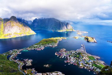 Göz kamaştırıcı dağların güneşli panoramik manzarası ve Reinebringen tepesinden Norveç 'in Lofoten adalarındaki Reine köyü. Gökkuşağı ve engebeli tepeleri olan manzaralı bir manzara.