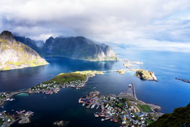 Göz kamaştırıcı dağların güneşli panoramik manzarası ve Reinebringen tepesinden Norveç 'in Lofoten adalarındaki Reine köyü. Gökkuşağı ve engebeli tepeleri olan manzaralı bir manzara.