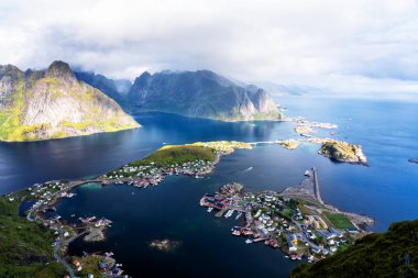 Göz kamaştırıcı dağların güneşli panoramik manzarası ve Reinebringen tepesinden Norveç 'in Lofoten adalarındaki Reine köyü. Gökkuşağı ve engebeli tepeleri olan manzaralı bir manzara.