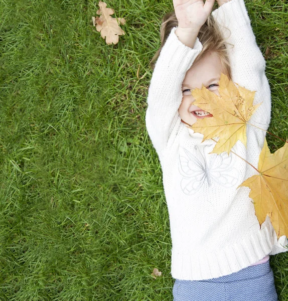 Веселая девушка лежит на зеленой траве, покрытой желтыми листьями клена — стоковое фото