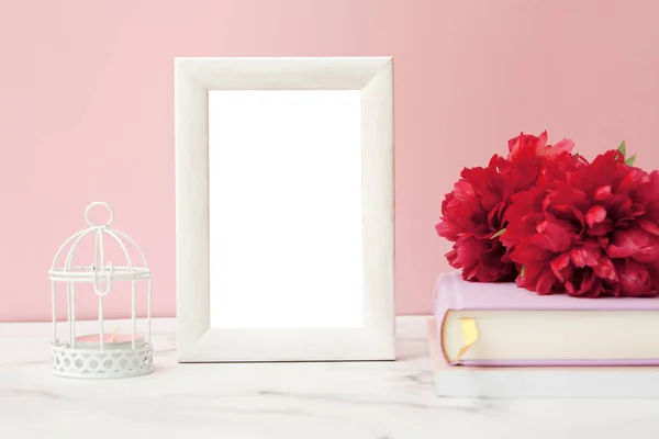 Mock-up white photo frame on the table next to flowers, books and a candlestick on a pink wall