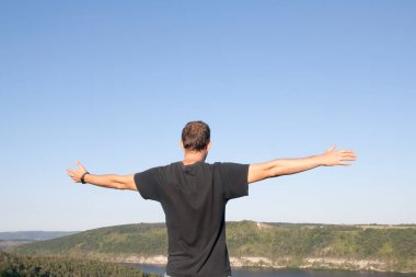 Kollarını gökyüzünün ve doğanın arka planına doğru uzatmış bir adam. İyileşme, keyif, denge konsepti